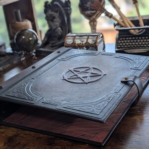 May include: A dark gray leather-bound book with a raised pentagram design on the cover. The book has decorative corner accents and a leather strap closure. The pages are a reddish-brown color.