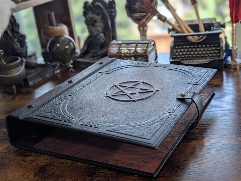May include: A dark gray leather-bound book with a raised pentagram design on the cover. The book has decorative corner accents and a leather strap closure. The pages are a reddish-brown color.