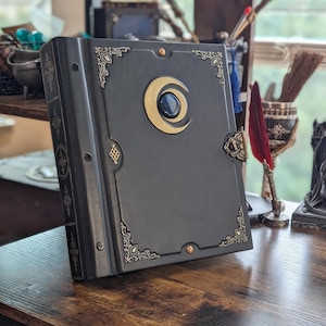 May include: A dark grey leather-bound book with gold-coloured detailing. The cover features a crescent moon design with a blue stone centre. Decorative corner pieces and a clasp enhance the book's ornate appearance. The book is on a wooden surface.