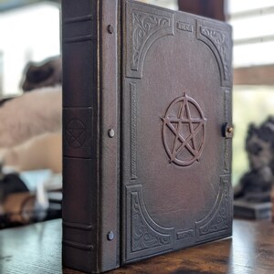 May include: A dark brown, leather-bound book with a pentagram emblem and ornate corner designs. The book has a clasp closure and is standing upright on a wooden surface. The spine has a series of rivets.
