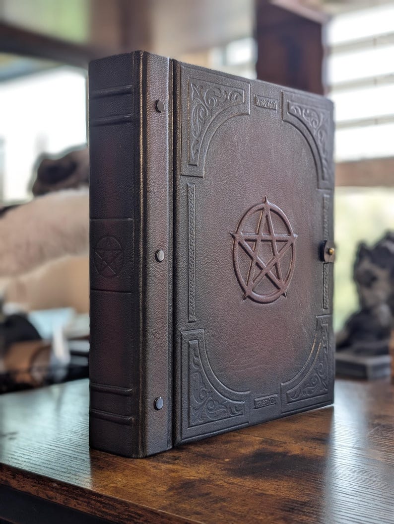 May include: A dark brown, leather-bound book with a pentagram emblem and ornate corner designs. The book has a clasp closure and is standing upright on a wooden surface. The spine has a series of rivets.
