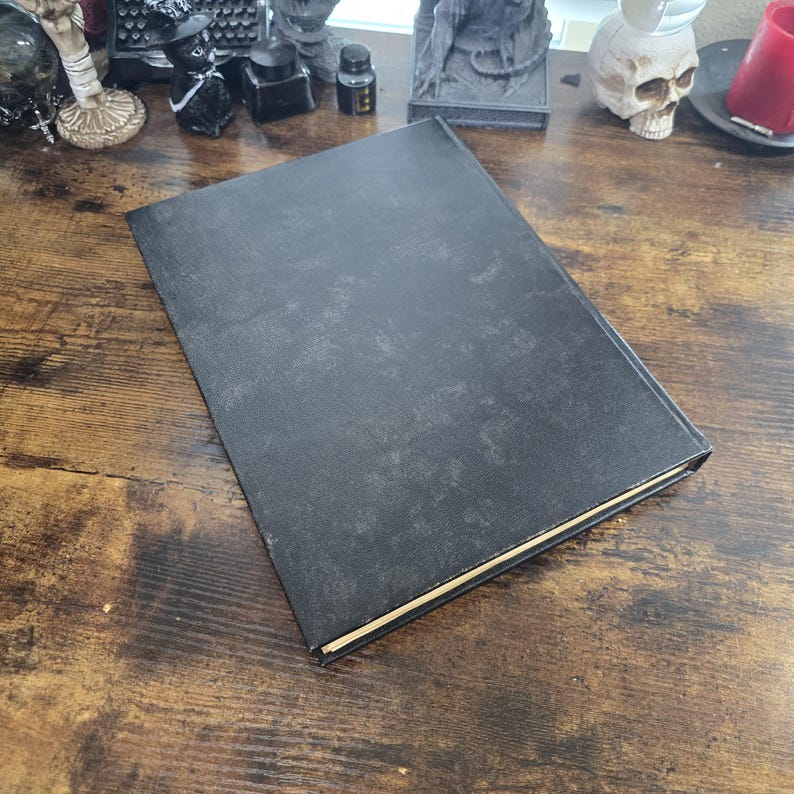 K&ouml;nnte beinhalten: Ein schwarzes Hardcover-Buch mit einer strukturierten Oberfl&auml;che. Das Buch liegt geschlossen auf einer Holzoberfl&auml;che.