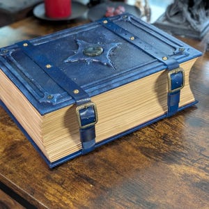 May include: A large, blue, leather-bound book with a starburst design on the cover. The book has brass buckles and straps. The pages are aged and yellowed, giving it a vintage look. The book is sitting on a wooden surface.