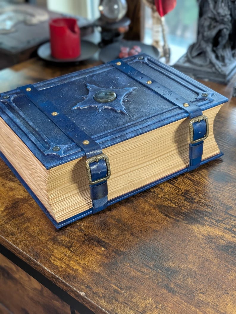 May include: A large, blue, leather-bound book with a starburst design on the cover. The book has brass buckles and straps. The pages are aged and yellowed, giving it a vintage look. The book is sitting on a wooden surface.