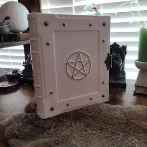 May include: White leather-bound journal with a pentagram design embossed on the front cover. The journal has brass accents and is closed.