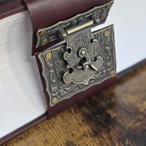 May include: A close-up of a brown leather-bound book with a decorative metal clasp. The clasp is ornate and has a floral design. The book is closed and lying on a wooden surface.