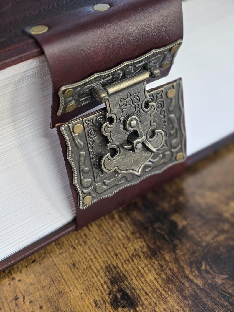 May include: A close-up of a brown leather-bound book with a decorative metal clasp. The clasp is ornate and has a floral design. The book is closed and lying on a wooden surface.
