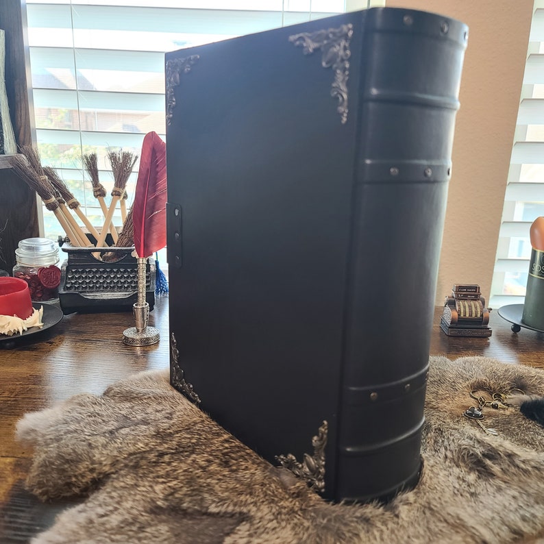 May include: A large, black book with ornate silver corner accents and a riveted spine. The book is closed and appears to be leather-bound. The book is on a fur-like surface.