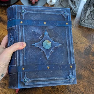 May include: A blue leather-bound book with a celestial design. The cover features a central starburst with a blue-green stone, accented by brass studs and straps. The book has a textured, speckled appearance.