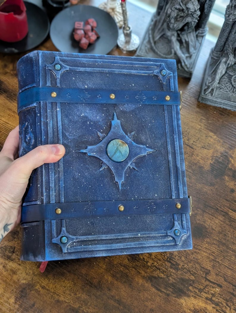 May include: A blue leather-bound book with a celestial design. The cover features a central starburst with a blue-green stone, accented by brass studs and straps. The book has a textured, speckled appearance.