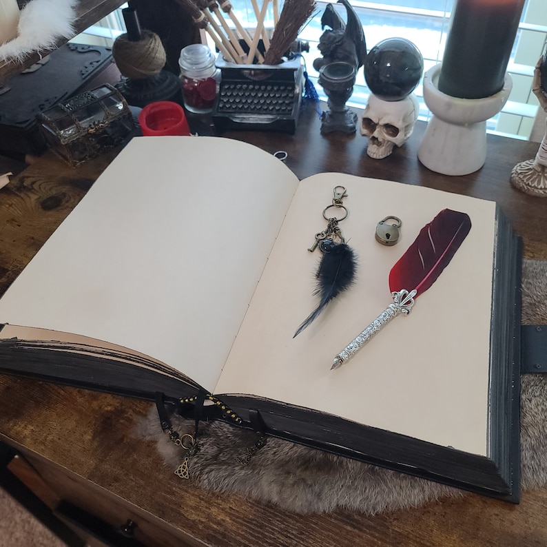 May include: An open journal with a black cover, a decorative feather pen, a small padlock, and a keychain with a black feather and keys. The journal is on a wooden surface, with gothic-themed items in the background, such as a skull.