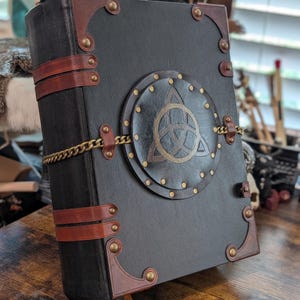 May include: A black leather-bound journal with brown leather accents and brass hardware. The cover features a circular emblem with a Celtic knot design and is secured with a gold chain. A brown ribbon bookmark is visible.