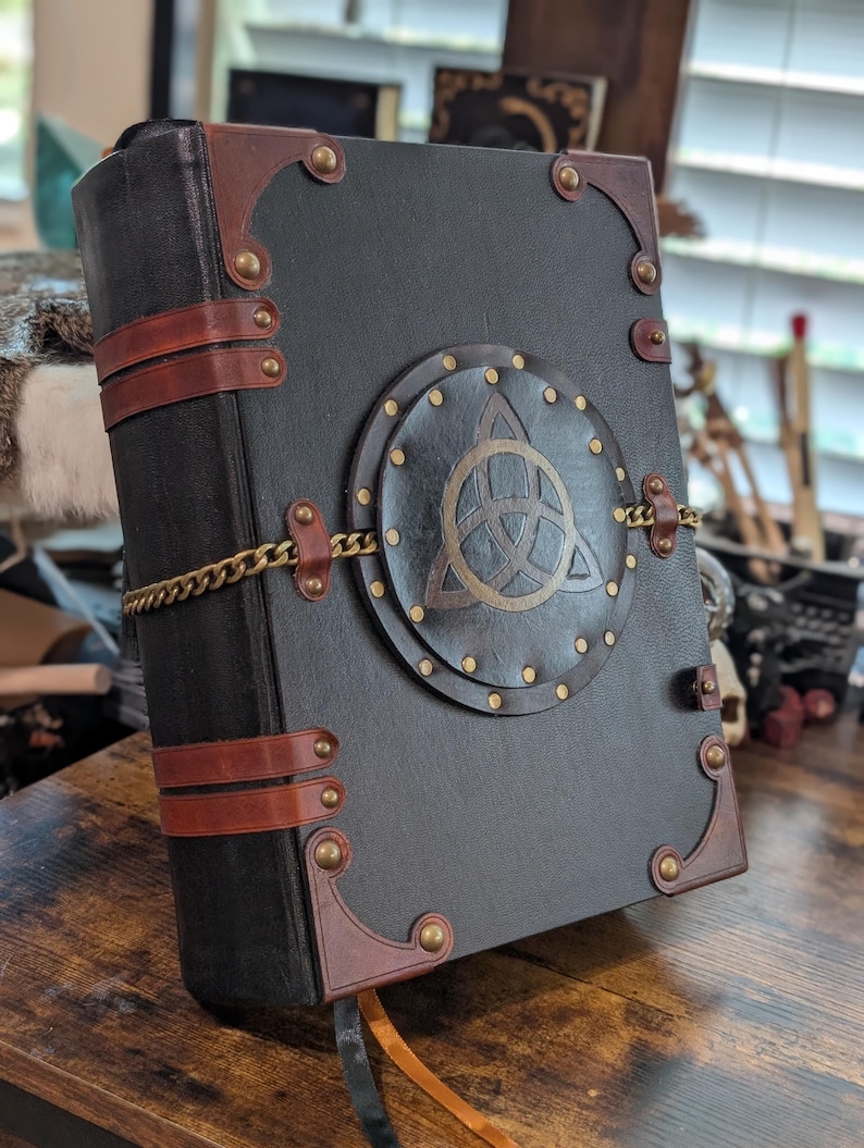 May include: A black leather-bound journal with brown leather accents and brass hardware. The cover features a circular emblem with a Celtic knot design and is secured with a gold chain. A brown ribbon bookmark is visible.