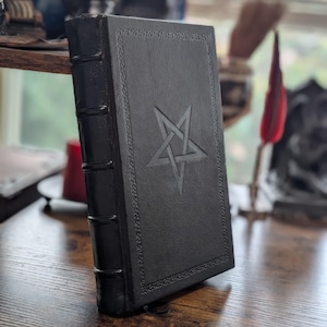 May include: A black leather-bound book with a pentagram embossed on the cover. The book has decorative border details and a ribbon bookmark. The spine has three raised bands. The book is on a wooden surface.