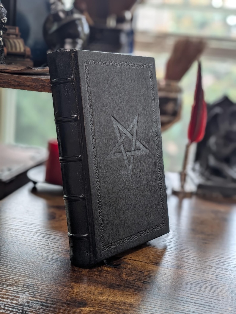 May include: A black leather-bound book with a pentagram embossed on the cover. The book has decorative border details and a ribbon bookmark. The spine has three raised bands. The book is on a wooden surface.