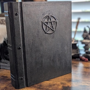 May include: A black book with a textured cover featuring an embossed pentagram symbol in the center. The spine is reinforced with a vertical strip secured by three visible rivets, and the front cover is framed by a thin, debossed rectangular border.