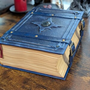 May include: A blue leather-bound book with a decorative clasp and a gemstone centerpiece. The book has a detailed cover design with metal accents and a red ribbon bookmark. The pages are aged, giving it a vintage look.