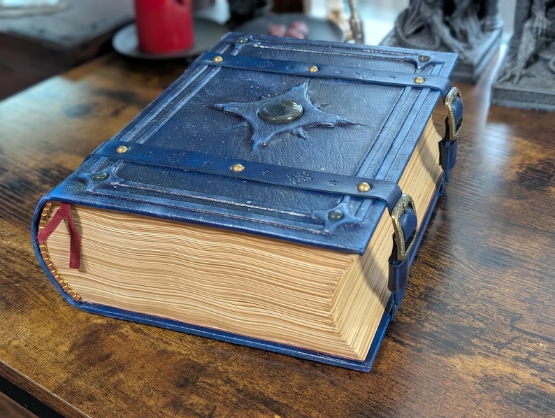 May include: A blue leather-bound book with a decorative clasp and a gemstone centerpiece. The book has a detailed cover design with metal accents and a red ribbon bookmark. The pages are aged, giving it a vintage look.