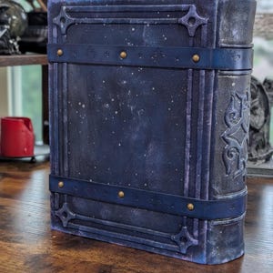 May include: A large, dark blue book with a celestial design. The cover features a textured, speckled finish with decorative straps and brass-colored rivets. The spine has an embossed pattern. The book is standing upright on a wooden surface.