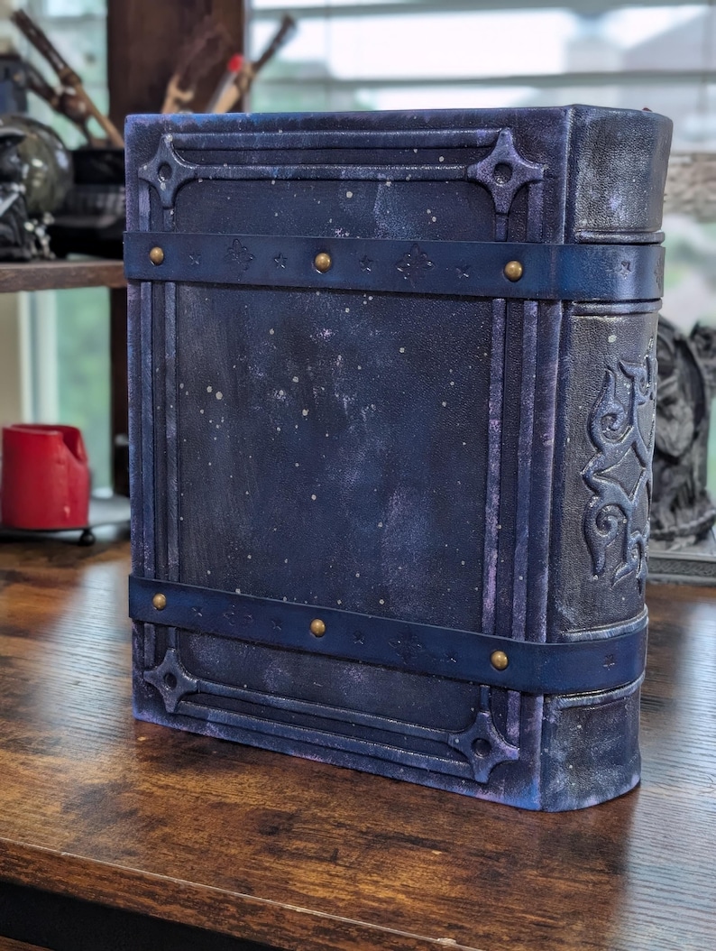 May include: A large, dark blue book with a celestial design. The cover features a textured, speckled finish with decorative straps and brass-colored rivets. The spine has an embossed pattern. The book is standing upright on a wooden surface.