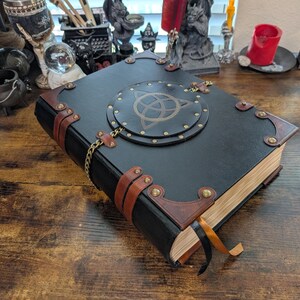 May include: A black leather-bound book with brown leather accents and brass hardware. The cover features a circular design with a gold symbol. A gold chain and ribbon bookmark add detail to this fantasy-themed journal.
