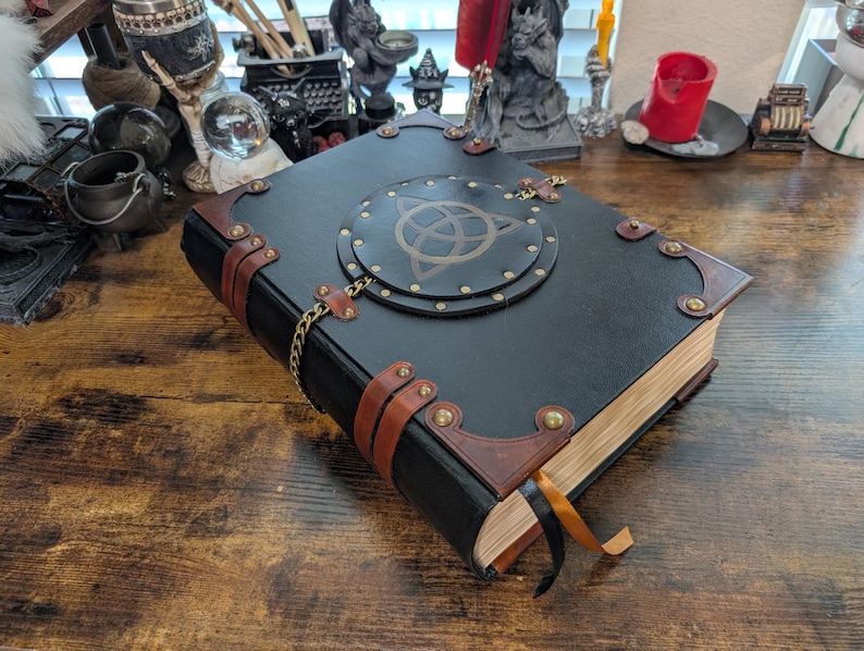 May include: A black leather-bound book with brown leather accents and brass hardware. The cover features a circular design with a gold symbol. A gold chain and ribbon bookmark add detail to this fantasy-themed journal.