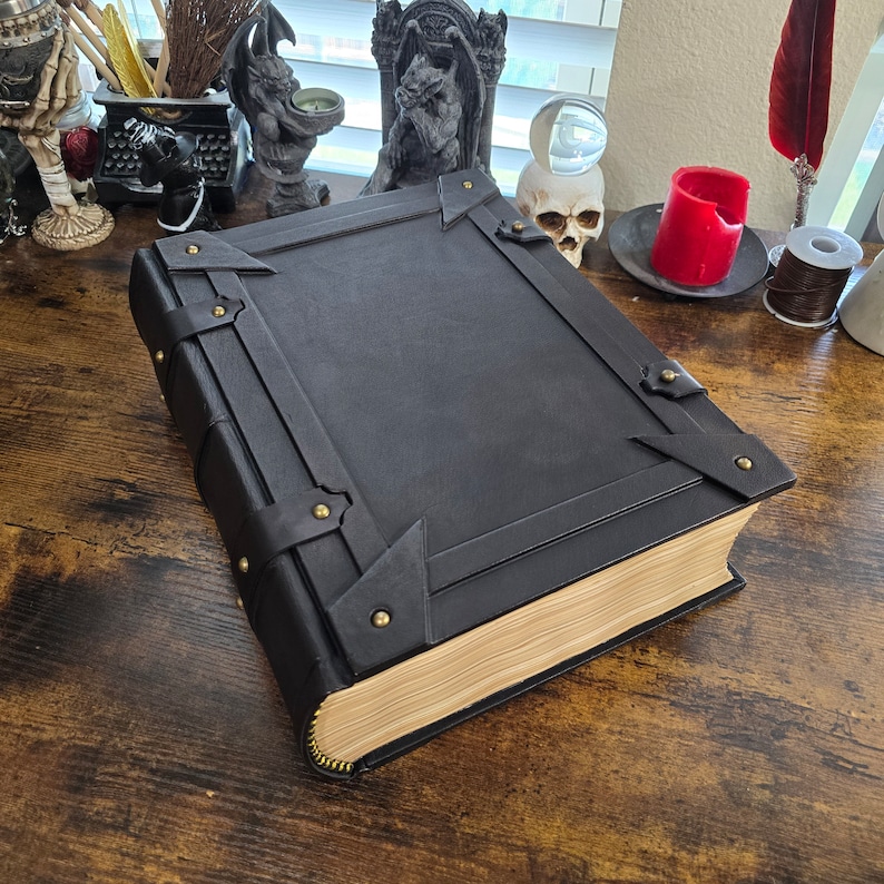 May include: A large, black leather-bound book with decorative metal accents and a thick spine. The book is closed and lying on a wooden surface.