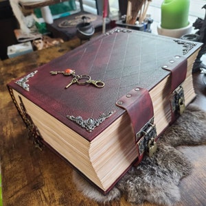 K&ouml;nnte beinhalten: Ein gro&szlig;es, burgunderfarbenes, ledergebundenes Buch mit verzierten Metallapplikationen und einem Schl&uuml;ssel mit einem roten Juwel. Das Buch ist geschlossen und hat einen dicken Stapel Seiten.
