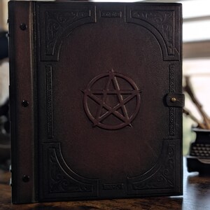 May include: A dark brown leather-bound book with a raised pentagram design in the center. The cover features embossed decorative patterns and a brass clasp. The book's spine has three rivets.