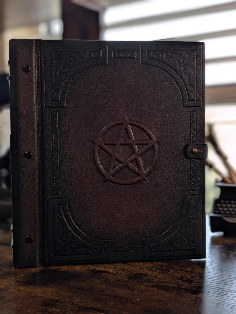 May include: A dark brown leather-bound book with a raised pentagram design in the center. The cover features embossed decorative patterns and a brass clasp. The book's spine has three rivets.
