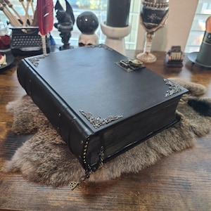May include: A large, black leather-bound book with decorative metal corner accents and a clasp closure. The book has a metal bookmark with a Celtic knot charm. The book is on a faux fur surface.