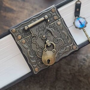 May include: A black leather-bound journal with a decorative metal clasp and a key with a blue glass eye charm. The journal has a white interior with pages.