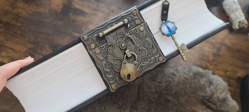 May include: A black leather-bound journal with a decorative metal clasp and a key with a blue glass eye charm. The journal has a white interior with pages.