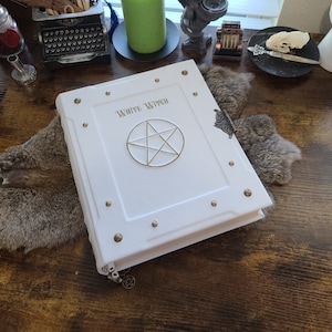 May include: White leather-bound journal with a gold pentagram and the words "White Witch" embossed on the cover. The journal has decorative metal accents and a clasp.