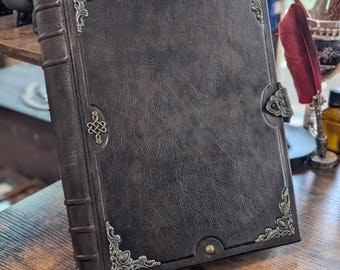 May include: A dark brown leather-bound journal featuring ornate antique brass-toned metal corner protectors, a central Celtic knot emblem, and a side latch closure. The spine has raised bands, and the cover is secured with small brass rivets.