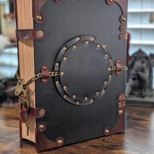 May include: A large, leather-bound book with a black cover and brown leather accents. The book features brass rivets, corner protectors, and a decorative circular element. A chain and lock secure the book, with a key charm.