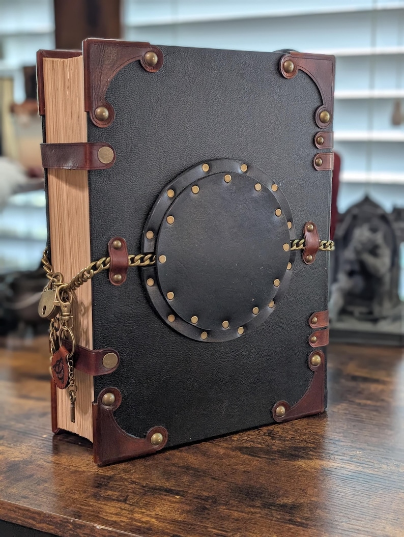 May include: A large, leather-bound book with a black cover and brown leather accents. The book features brass rivets, corner protectors, and a decorative circular element. A chain and lock secure the book, with a key charm.