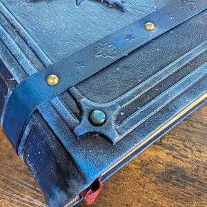 May include: Close-up of a blue leather-bound journal with embossed designs and a decorative strap. The journal features a gemstone accent and gold-colored rivets. A red ribbon bookmark is visible.
