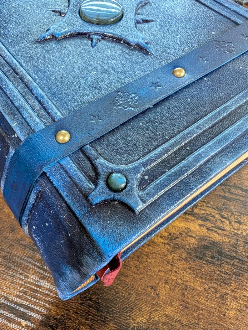 May include: Close-up of a blue leather-bound journal with embossed designs and a decorative strap. The journal features a gemstone accent and gold-colored rivets. A red ribbon bookmark is visible.