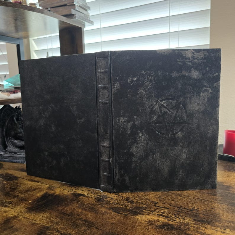 K&ouml;nnte beinhalten: Ein schwarzes, ledergebundenes Buch mit einem gepr&auml;gten Pentagramm auf dem vorderen Einband. Das Buch hat ein abgenutztes, altes Aussehen und einen R&uuml;cken mit erhabenen B&auml;ndern.