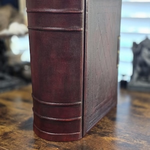 May include: A dark brown leather-bound book with a decorative embossed pattern on the cover. The book is closed and standing upright on a wooden surface.