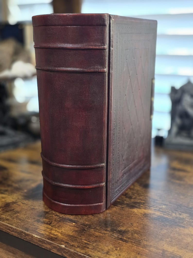 May include: A dark brown leather-bound book with a decorative embossed pattern on the cover. The book is closed and standing upright on a wooden surface.