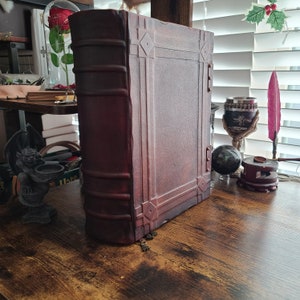 K&ouml;nnte beinhalten: Ein gro&szlig;es, dunkelbraunes, ledergebundenes Buch mit aufwendigen Pr&auml;gungen und Metallverzierungen. Das Buch ist geschlossen und liegt auf einer Holzoberfl&auml;che.