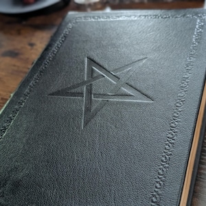 May include: A dark green leather-bound book with a pentagram embossed on the cover. The cover has a decorative border and the pages have a gold edge. The book is closed and resting on a wooden surface.