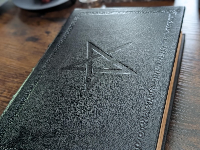 May include: A dark green leather-bound book with a pentagram embossed on the cover. The cover has a decorative border and the pages have a gold edge. The book is closed and resting on a wooden surface.
