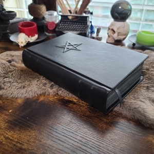 May include: A black leather-bound book with a silver pentagram emblem on the cover. The book has a black ribbon bookmark and is resting on a faux fur surface. The background includes a skull, candles, and a typewriter.