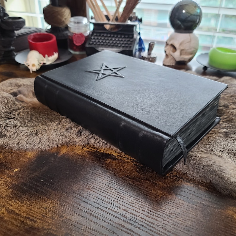 May include: A black leather-bound book with a silver pentagram emblem on the cover. The book has a black ribbon bookmark and is resting on a faux fur surface. The background includes a skull, candles, and a typewriter.