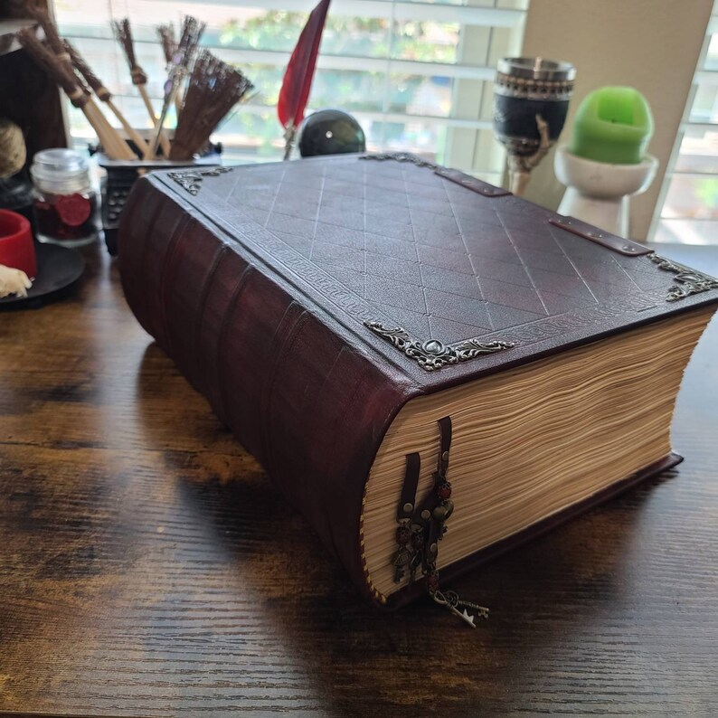 K&ouml;nnte beinhalten: Ein gro&szlig;es, dunkelbraunes, ledergebundenes Buch mit verzierten Metallverzierungen und einem dekorativen Quasten. Das Buch ist geschlossen und liegt auf einer Holzoberfl&auml;che.