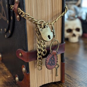 May include: A close-up of a leather-bound book with a brass lock and chain. The book has a brown leather strap and a key attached to a leather tag. The book's pages are aged, and the cover is black.