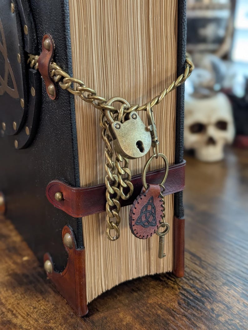 May include: A close-up of a leather-bound book with a brass lock and chain. The book has a brown leather strap and a key attached to a leather tag. The book's pages are aged, and the cover is black.